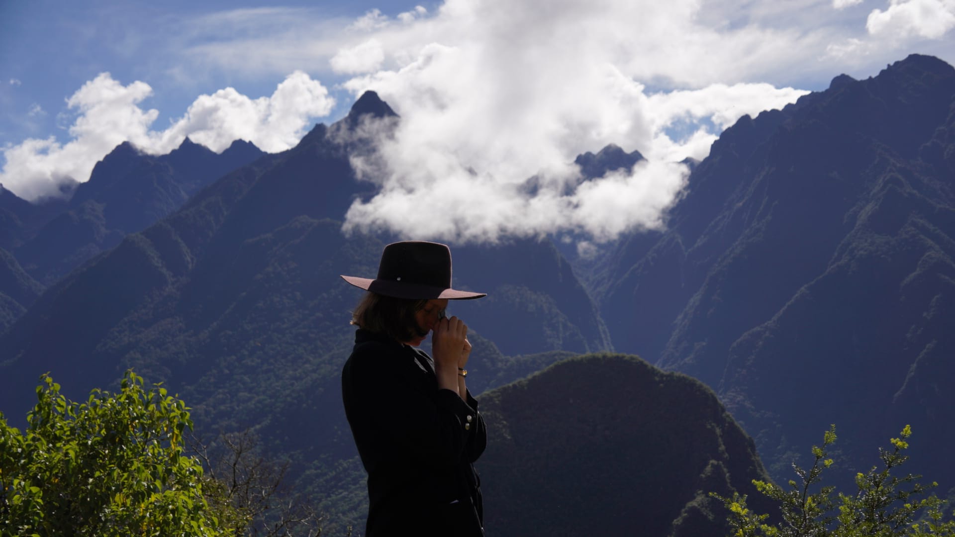 Agni Vedaa w kapeluszu, z dłońmi złożonymi przy twarzy, na górnych tarasach Machu Picchu, z widokiem na górę Yanantin w chmurach